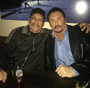Two men are sitting together at a table in a dimly lit bar. The man on the left has a mustache, wearing a black cap and a black zip-up jacket. The man on the right has a groomed mustache and is dressed in a dark leather jacket over a light blue dress shirt. They are both smiling and leaning towards each other, with drinks on the table in front of them. The background features a small, illuminated area with a photo or artwork on the wall.