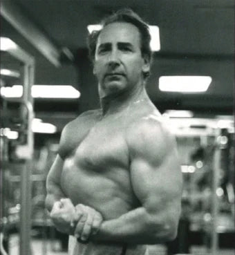 A muscular man is posing in a gym setting, showcasing his arm and shoulder muscles. He has a serious expression and is looking directly at the camera. The photo is in black and white, with various gym equipment visible in the background, including weights and machines. The lighting is bright, highlighting the definition of his muscles.