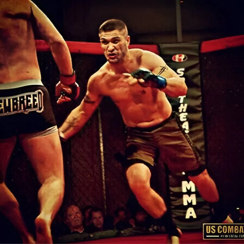 In a mixed martial arts (MMA) fight setting, two shirtless fighters are engaged in a competitive moment inside a cage. One fighter, on the left, is facing away from the camera, revealing his muscular back and wearing navy blue shorts. The other fighter, positioned on the right, has a determined expression and is lunging forward, wearing brown shorts and striking with his right fist. The background features a blurred crowd watching the match, and the cage has red and black padding with promotional signage.