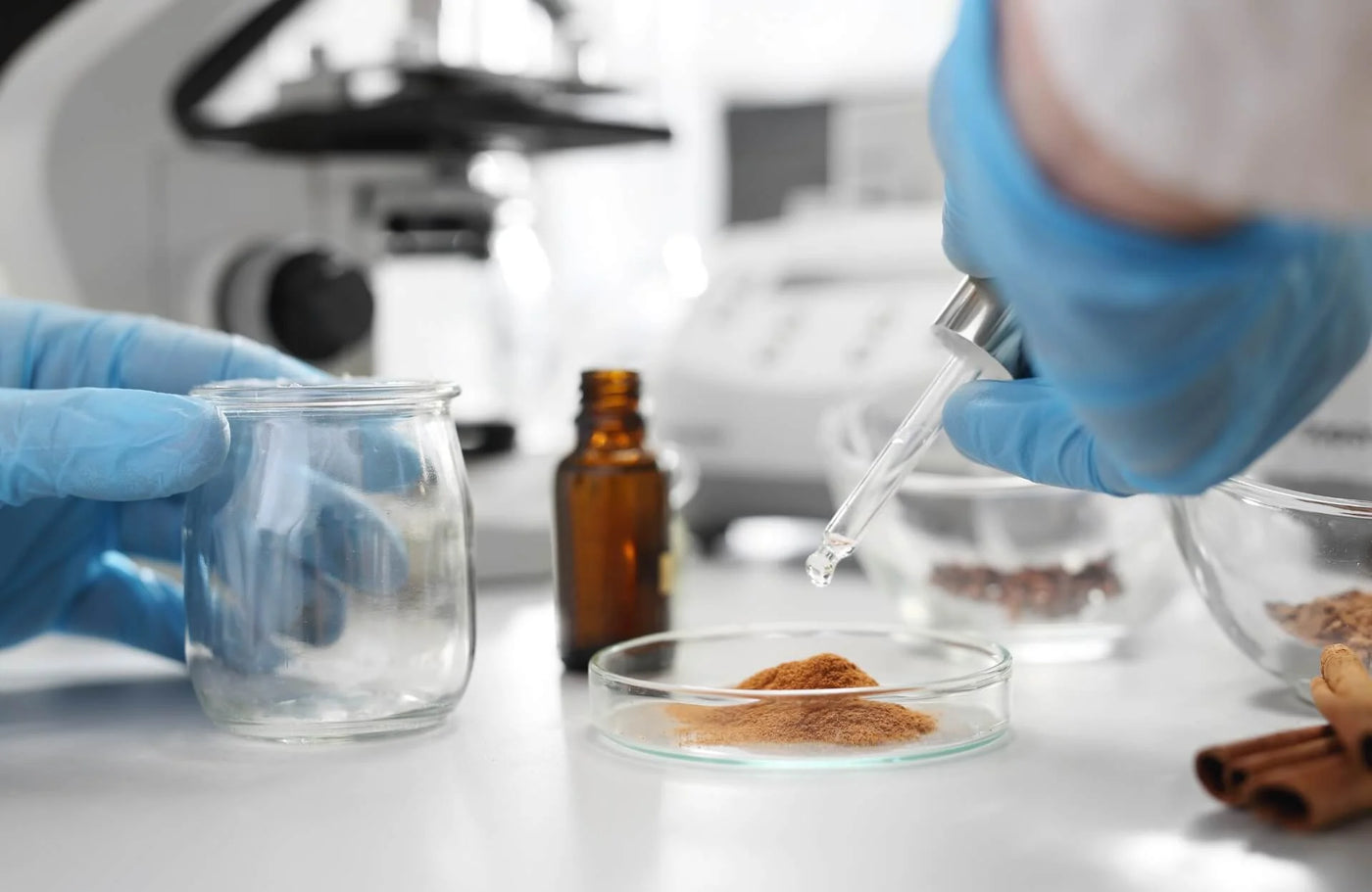 A pair of gloved hands are shown working in a laboratory setting. One hand holds a clear glass jar, while the other is using a dropper to dispense a liquid into a shallow glass dish containing brown powder. Nearby, there are glass containers with various ingredients, including a small amber bottle and a bowl with more powder and small brown items resembling seeds or grains. In the background, a scientific microscope and other lab equipment are faintly visible. The workspace is tidy and well-lit.