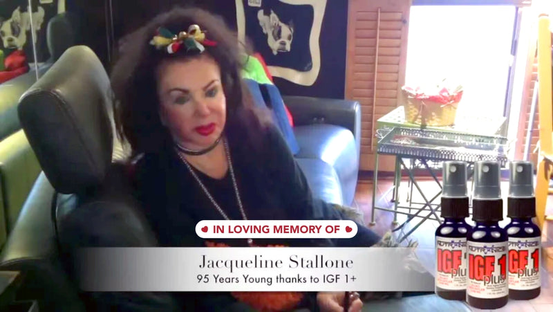 A woman with voluminous dark hair sits on a black leather couch, wearing a black top and layered necklaces. She has bright lipstick and a colorful hair accessory. To the left, there is a reflection of a dog's portrait on the wall. In front of her, a white banner reads "In Loving Memory Of" and below that, "Jacqueline Stallone, 95 Years Young thanks to IGF 1+" is displayed. To the right, there are four small spray bottles labeled "IGF 1 plus" on a table.
