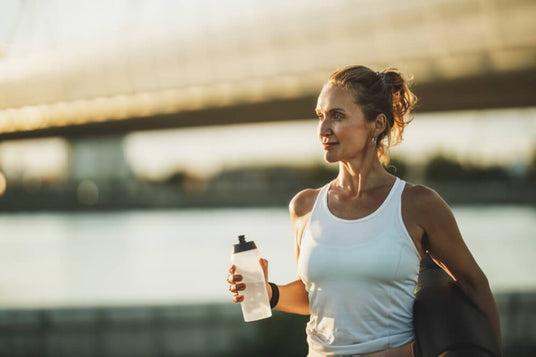 A middle-aged woman stands outdoors, holding a clear water bottle in one hand and a yoga mat rolled up in the other. She is wearing a white athletic tank top and has her hair pulled back in a bun. The background features a blurred bridge and water, with warm sunlight creating a golden hour effect. The woman appears relaxed and focused.