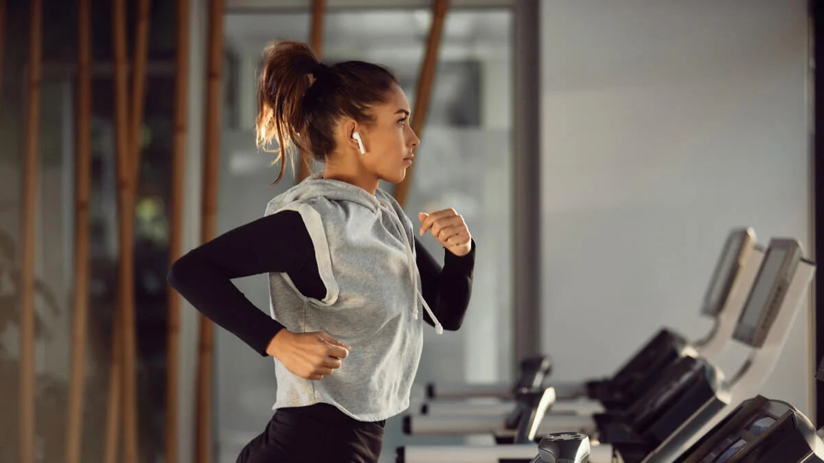 A young woman with long hair tied in a ponytail is jogging on a treadmill in a modern gym. She is wearing a light gray sleeveless hoodie over a black long-sleeve shirt and appears focused as she runs. White wireless earbuds are in her ears, and there are exercise machines in the background, indicating an active fitness environment. Natural light is coming through large windows, and there are wooden elements in the design of the gym.
