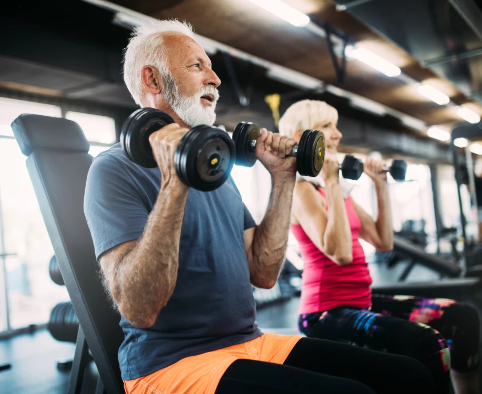 An elderly man with a white beard is sitting on a weight bench, lifting two dumbbells at shoulder height. He is wearing a blue t-shirt and orange shorts. Next to him, a woman with short, light-colored hair in a pink tank top is also lifting dumbbells. Both are focused on their workout in a gym setting, surrounded by various exercise equipment and bright overhead lights.