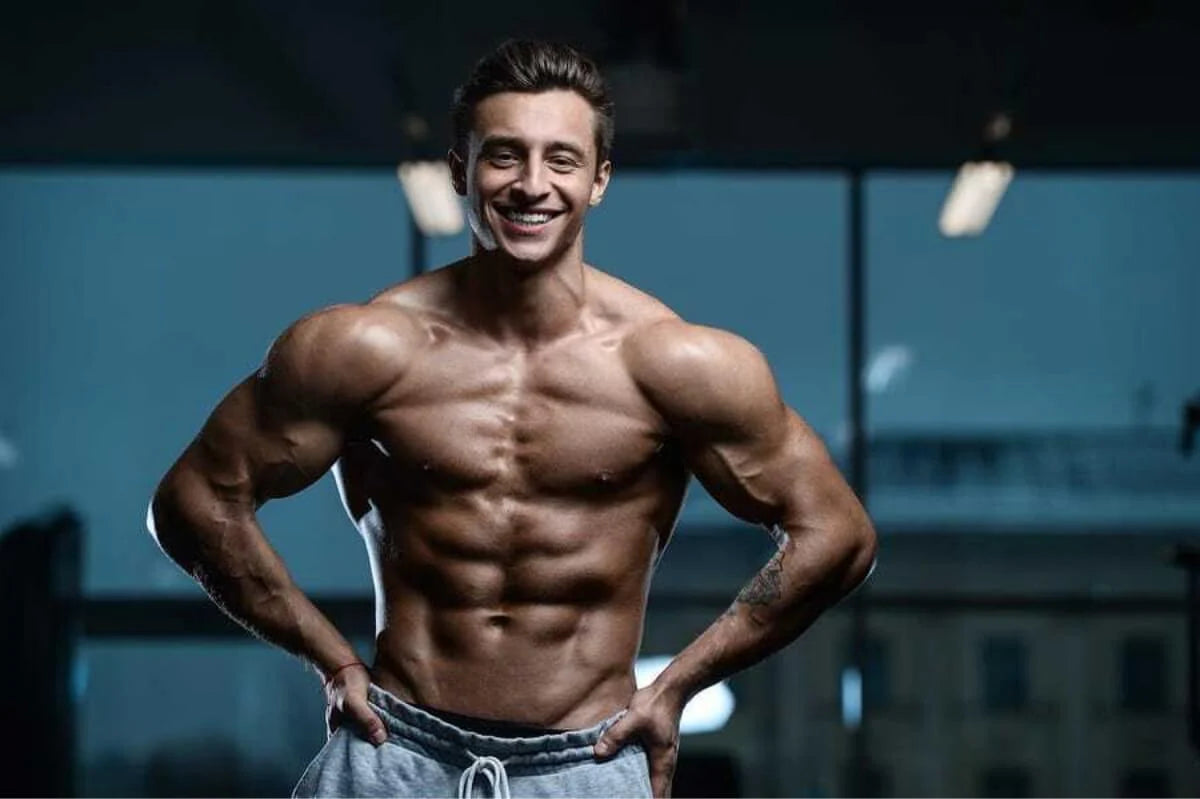 A muscular man is posing in a fitness studio with a large smile, showcasing his toned physique. He has prominent arm and chest muscles with defined abs. He is wearing gray sweatpants and standing confidently with his hands resting on his hips. The background features soft lighting and hints of gym equipment, creating a fit and energetic atmosphere.