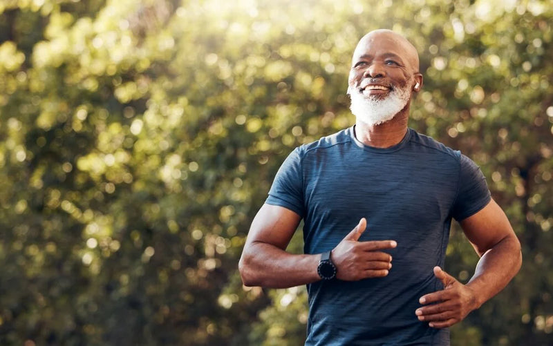 A smiling older man with a salt-and-pepper beard is jogging outdoors. He is wearing a dark blue athletic t-shirt and a smartwatch on his left wrist. The background features blurred green foliage, suggesting a sunny day in a park or natural setting. The man appears to be enjoying his run, capturing a moment of fitness and vitality.