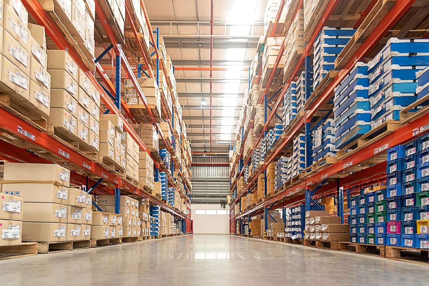 An interior view of a large warehouse filled with shelves stacked high with boxes and containers. The shelves are a mix of orange metal supports with varying sized cardboard boxes primarily stacked on wooden pallets. The boxes are labeled with white shipping labels displaying barcodes and descriptions. Some shelves are also filled with colorful plastic containers, predominantly blue, green, and yellow. The warehouse floor is smooth and reflective, with bright overhead lighting illuminating the space.