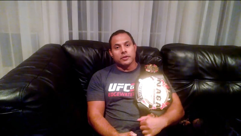 A man sits on a black leather couch, wearing a dark t-shirt with "UFC EDGEWATER" printed on it. He has a serious expression and is casually leaning back. Over his right shoulder, he holds a championship belt, which features symbols and logos. In the background, there are sheer curtains that suggest a well-lit room, creating a soft atmosphere.