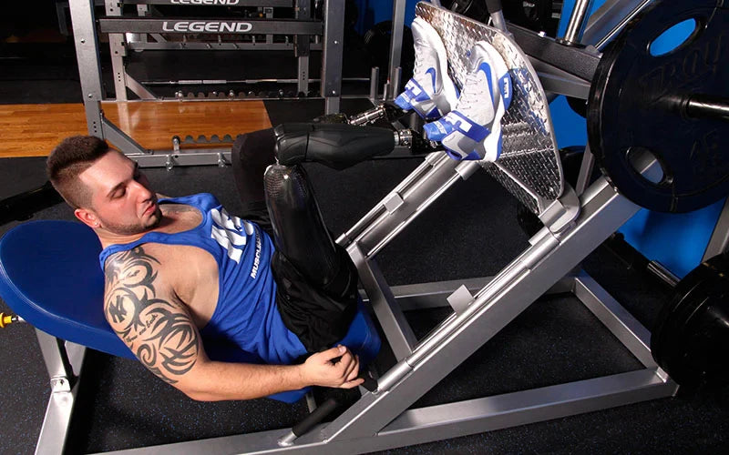 A male athlete with short dark hair and a visible tattoo on his right arm is using a leg press machine in a gym. He is wearing a blue tank top and black workout shorts. The machine is designed for leg exercises, and the athlete has prosthetic legs with blue and white shoes. The background shows gym equipment and a blue wall. The focus is on the athlete's determination and strength as he engages in his workout.