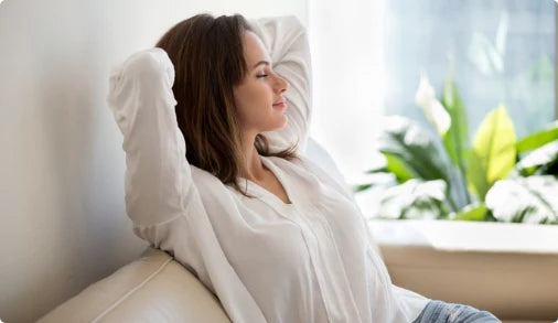 A woman with long, dark hair sits comfortably on a light-colored sofa, leaning back with her arms resting behind her head. She wears a loose-fitting white blouse and light jeans. Her eyes are closed, and she appears relaxed and at peace. In the background, there are green plants visible, adding a serene touch to the setting. Soft natural light filters in, creating a calm atmosphere.