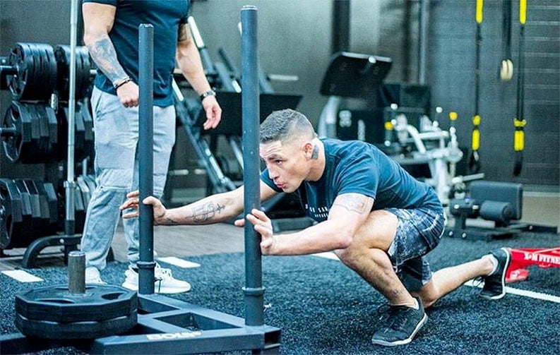 A man in a blue shirt and patterned shorts kneels on a gym floor while pushing a weighted sled. He has a focused expression as he leans forward, using both hands to grip the sled's poles. In the background, another individual in a black shirt and gray pants stands nearby, observing. The gym features various fitness equipment, including weights and resistance bands, with dark flooring and a modern setup.