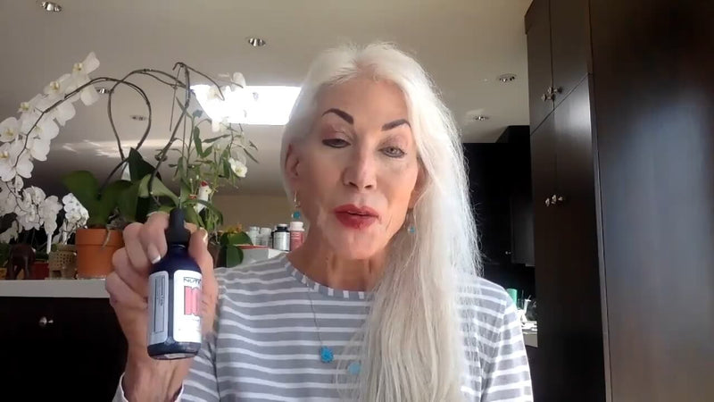 A woman with long, straight white hair and noticeable makeup is sitting at a table in a bright kitchen. She is wearing a striped long-sleeve shirt and a necklace with a blue pendant. In her right hand, she holds a small dark blue glass bottle with a dropper top, displaying a label. Behind her, there are several potted plants, including orchids, and a countertop with various containers and products visible. The space has modern dark cabinetry and overhead lighting.