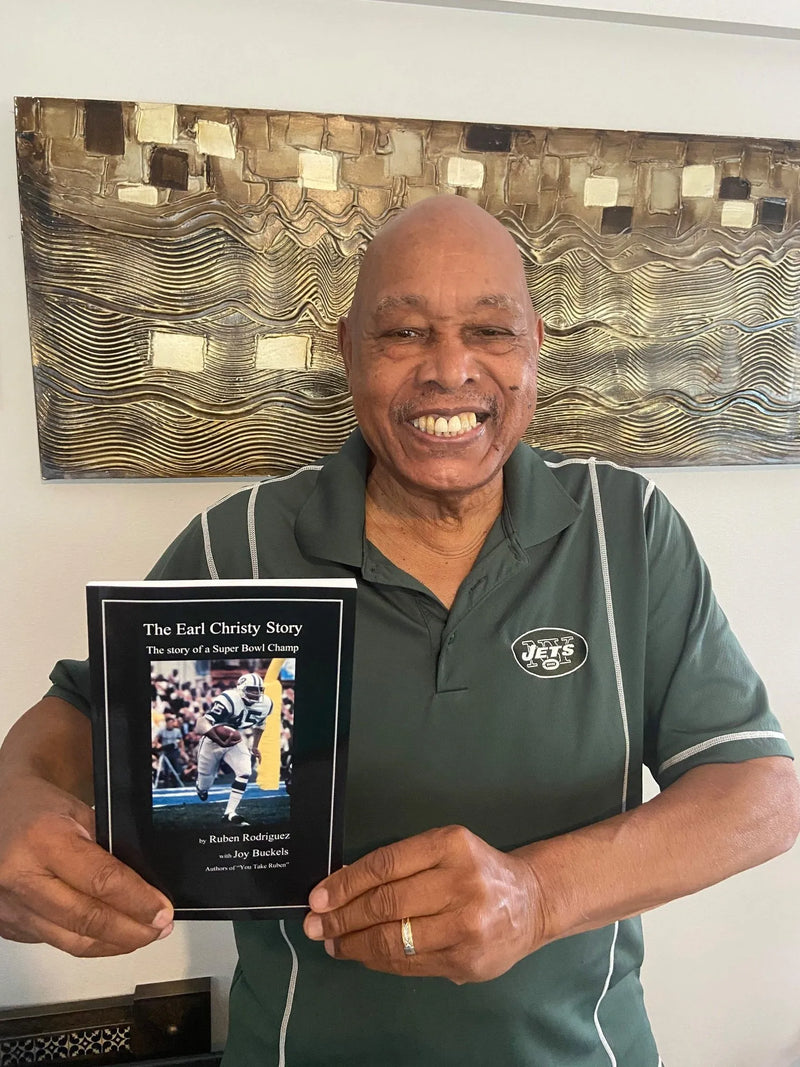 A smiling older man with a bald head and a light brown complexion stands in front of an abstract artwork featuring wavy gold and brown patterns. He is wearing a green shirt with a white logo resembling the New York Jets. He holds a book titled "The Earl Christy Story," which tells the story of a Super Bowl champion. The cover features a photo of a football player in action, wearing a white jersey with the number 15. The man is displaying the book with both hands, looking directly at the camera.