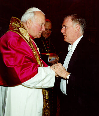 A historic moment showing Pope John Paul II in a white mitre and a red and gold ceremonial robe, warmly engaging in conversation while holding the hands of a man in a black tuxedo. Behind them, a man in a clerical collar, possibly a cardinal, observes the interaction. The background displays ornate, dark decorations typical of a formal chamber. The overall atmosphere conveys a sense of respect and camaraderie.