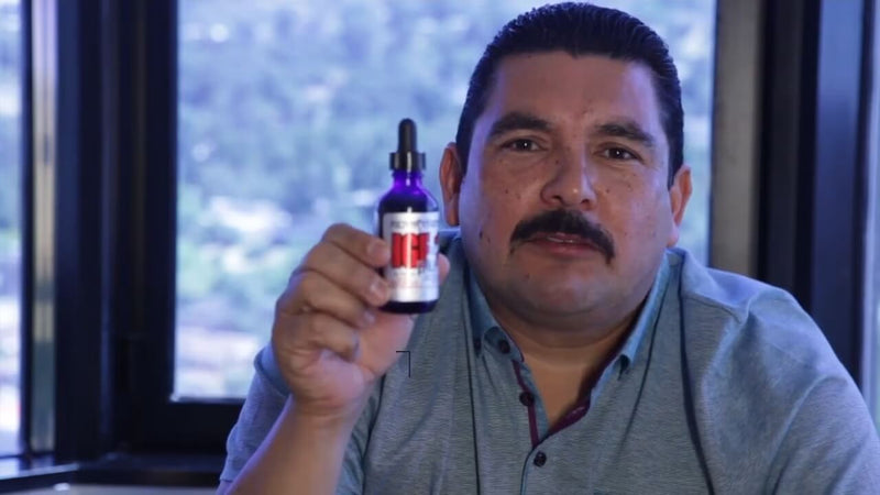 A man with a thick mustache is holding up a small bottle with a dropper, labeled in red and white, set against a background of large windows that reveal a blurred view of greenery and distant hills. He is wearing a short-sleeved light blue shirt with a subtle collar and has a focused expression on his face. The setting appears to be indoors with bright natural light illuminating the scene.