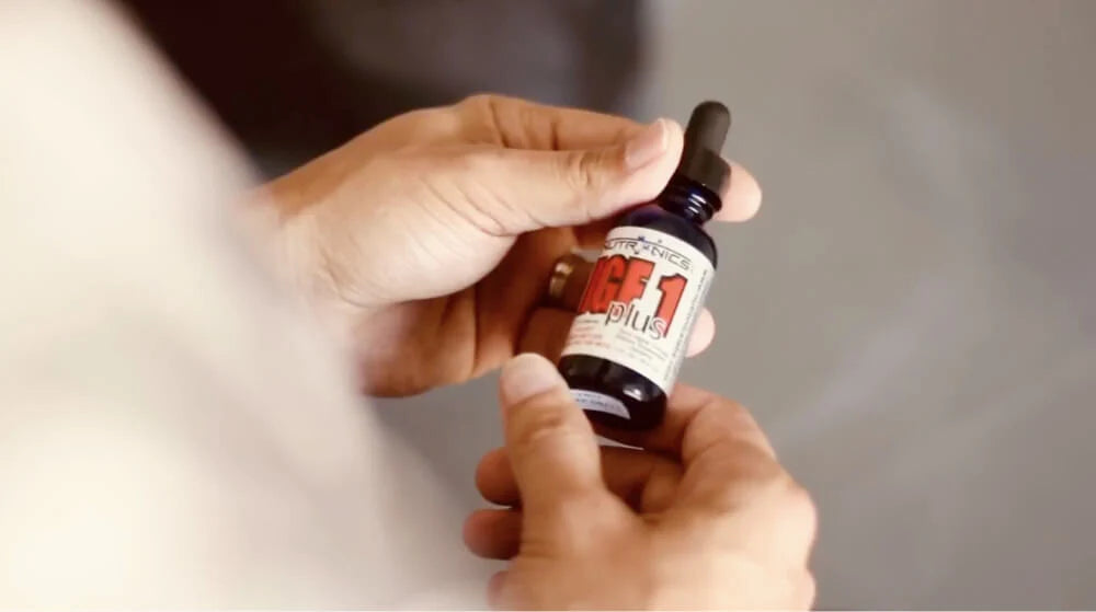A person is holding a small dark glass bottle with a dropper top. The bottle has a white and red label that reads "AGE1 plus" with additional text and graphics that are not clearly legible. The person's hands are visible, and they are positioned against a blurred background, suggesting an indoor setting.