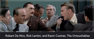 A group of men in 1920s attire are gathered in a tense scene. In the foreground, a man with slicked-back hair and a dark suit, possibly Kevin Costner, is animatedly speaking, gesturing with his hand toward another man who appears to be Robert De Niro, wearing a light-colored suit and a serious expression. Surrounding them are several other men, including one with a mustache and a cigarette. The setting suggests a confrontation or a critical conversation in a dimly lit room, typical of a dramatic moment.