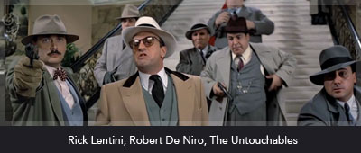A group of men dressed in 1920s gangster attire, featuring suits and fedoras, stand on a grand staircase. One man in the foreground, holding a gun, is wearing a light-colored overcoat and a white hat. The others appear stern, with serious expressions and also wear hats. The background suggests a lavish interior with decorative railings. The names Rick Lentini and Robert De Niro are mentioned along with the title The Untouchables at the bottom of the image.