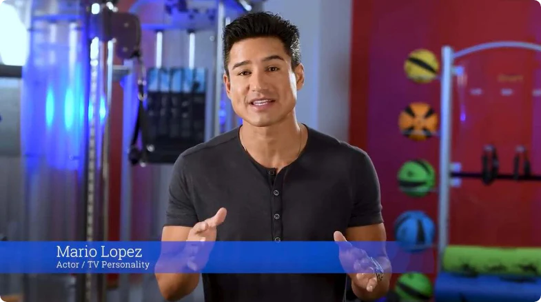 A man with dark hair and a fit physique is standing in a brightly lit room with exercise equipment in the background. He is wearing a short-sleeved black shirt and gesturing with his hands as he speaks. There is a blue banner at the bottom of the image displaying his name, Mario Lopez, along with the text Actor / TV Personality. The background features colorful exercise balls and gym apparatus.