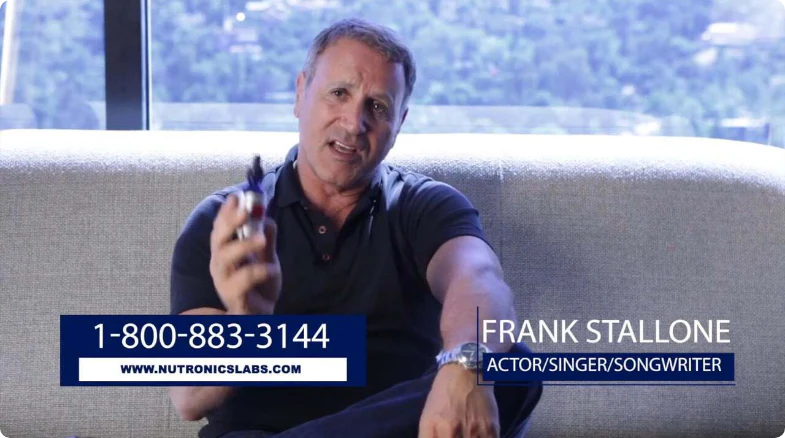 A middle-aged man with short, slightly wavy hair and a well-defined physique is sitting on a light-colored sofa. He is wearing a dark polo shirt and is holding a small object in his right hand. The background features a blurred view of greenery and hills through large windows. Text overlays at the bottom include a phone number, a website URL, and the name "Frank Stallone" with the titles "Actor/Singer/Songwriter" below it.