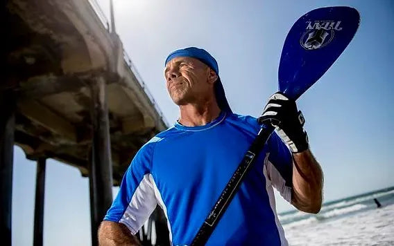 A man stands confidently on a beach, wearing a bright blue athletic shirt with white accents and a blue headband. He holds a paddle with a dark handle and a blue blade, gazing into the distance with a determined expression. In the background, a pier extends over the water, and gentle waves can be seen lapping at the shore. The scene is illuminated by sunlight, creating a vibrant and active atmosphere.